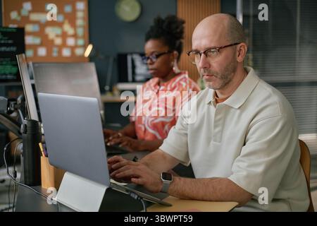 Middle aged Caucasian man wearing glasses working on laptop at desk with Black woman using computer in modern office setting, both focused on tasks, technology visible Stock Photo