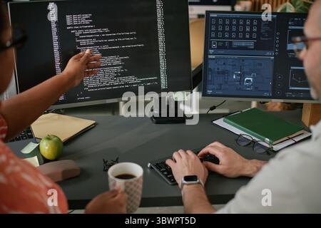 Caucasian middle aged man and Black woman collaborating at workstation, analyzing code on computer monitors, discussing software development, holding coffee mug, using keyboard Stock Photo