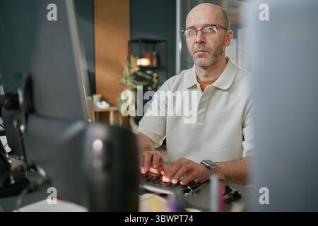 Portrait of middle aged Caucasian man wearing glasses working at computer in modern office, typing on keyboard and looking at monitor with focused expression Stock Photo