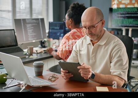 Middle aged Caucasian man using digital tablet at desk while Black woman working on computer in background, both focused on technology tasks in modern office workspace Stock Photo