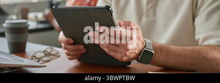 Website header shot of Caucasian middle aged man holding digital tablet with both hands sitting at desk near open laptop and eyeglasses, focusing on screen in office environment Stock Photo