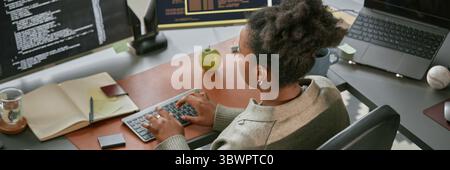 Website header shot of young adult Black woman working at computer desk, typing on keyboard and analyzing code on monitor, surrounded by office supplies and electronic devices in modern workspace Stock Photo