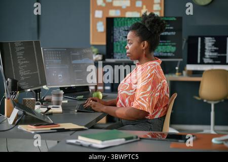 Young adult Black woman working at computer workstation in modern office, typing on keyboard and focusing on multiple monitors displaying programming code and data Stock Photo
