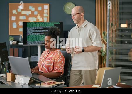 Black woman sitting at desk working on computer while middle aged Caucasian man standing beside her holding mug and watching screen in modern office environment Stock Photo