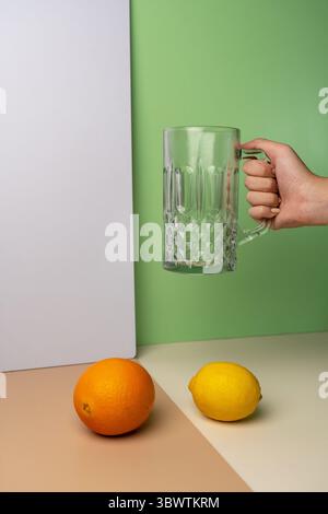 Hand holding an empty crystal-cut glass beer mug. An orange and a lemon sit below on a modern, multi-colored background with copy space. Stock Photo