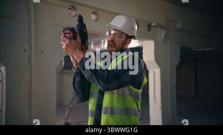 Angry Caucasian foreman male guy engineer man builder holding drill electric tool constructor drilling in factory construction worker doing repairment Stock Photo