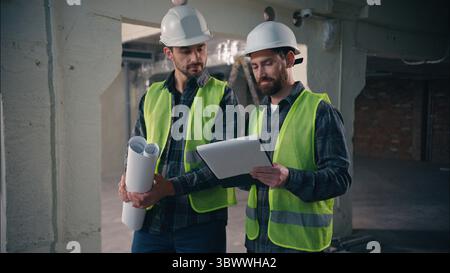 Caucasian two people men architects engineers guys males builders in helmets hardhats and vests discussing building plan project looking blueprint Stock Photo