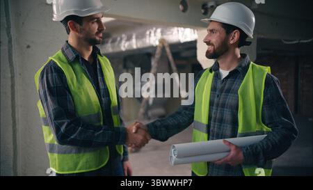 Satisfied Caucasian guys discussing draft handshake in construction contractors men technicians repairmen looking blueprint site colleagues builders Stock Photo