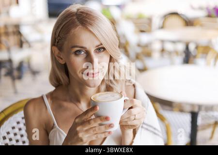 Portrait pretty blonde woman, smiling woman in cozy plaid, sitting in cafe with mug of hot coffee rejoices in a happy morning Stock Photo