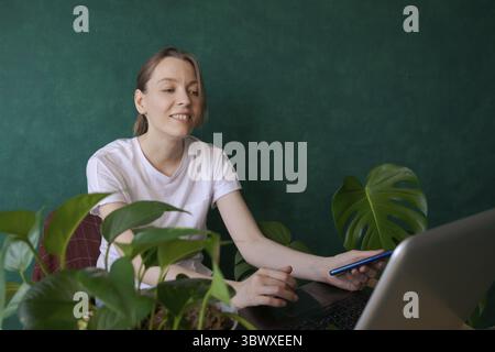 Beautiful young woman sits on chair at table with laptop next to an indoor green and works on a computer against a green wall. Work from home, blogger Stock Photo