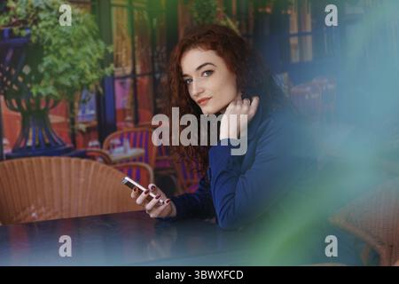 Portrait happy smiling pretty redhead girl sitting in street cafe or co-working space, using mobile phone, texting or watching live broadcast, online Stock Photo