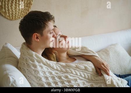 Dreamy married couple young people in knitted blanket on sofa, looking into distance, planning common future or remembering memories, enjoying peacefu Stock Photo