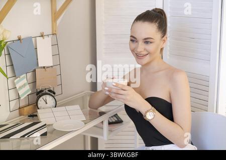 Happy beautiful young woman sitting at her desk at home with mug of coffee in her hand Stock Photo