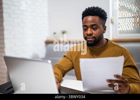 Young African businessman typing on laptop and analyzing documents in living room. He is working remotely from his home. Stock Photo