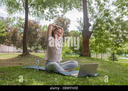 Smiling woman doing triceps stretch during outdoor yoga session with laptop in park setting. Stock Photo