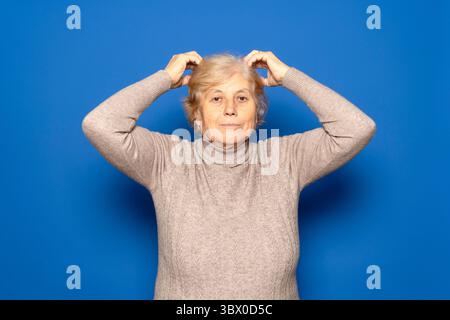 Caucasian woman in her 70s wearing a dark beige sweater scratching her head worriedly trying to remember something important, isolated over blue Stock Photo