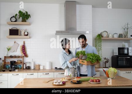 Happy young couple preparing fresh organic vegetables in their modern kitchen. Smiling man holds box of greens while woman points at lettuce, creating Stock Photo
