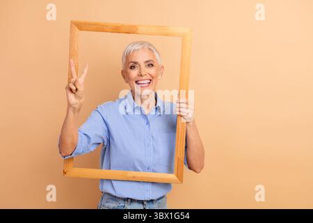Cheerful senior woman with short white hair holding a frame and making a peace sign in a casual outfit Stock Photo