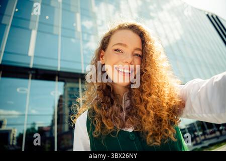 Confident curly-haired businesswoman poses outdoors with a stylish waistcoat against modern city buildings background. Stock Photo