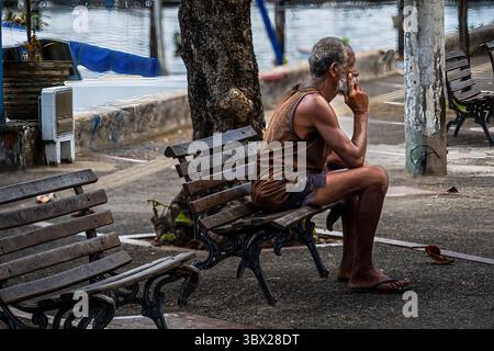 Salvador, Bahia, Brazil— March 10, 2025. A long distance telephoto image of a man thinking while sitting on a park bench. Stock Photo