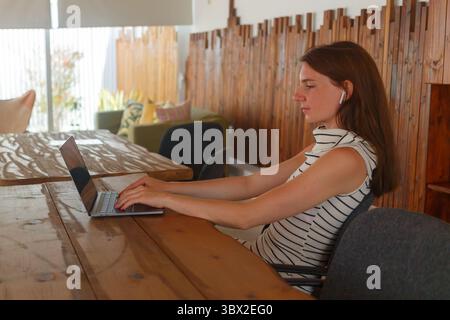 A young woman is focused on her laptop in a cozy wooden cafe, surrounded by natural light and stylish decor, during a sunny afternoon. Stock Photo