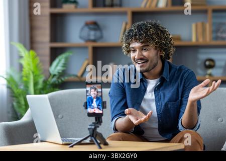A young man with curly hair is video chatting on his phone while using a laptop at home, gesturing with his hands. Stock Photo