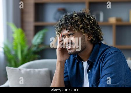 A young man with curly hair experiencing a headache, sitting indoors and holding his head in pain. Stock Photo