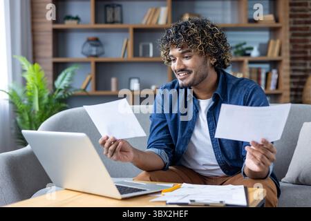 A young man reviews documents while using his laptop in a comfortable home setting, smiling and engaged with his work. Stock Photo
