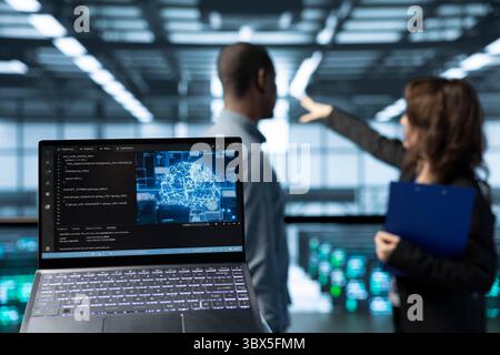 Laptop in front of data center technicians in blurry background deploying AI driven management tools, inspecting server clusters, updating network topologies, validating storage integrity Stock Photo