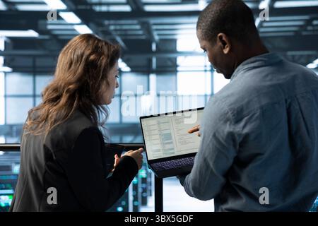 Programmers in data center workspace coding on laptop, analyzing metrics, optimizing storage volumes. Teamworking IT professionals in server hub using notebook, chatting, ensuring seamless operations Stock Photo