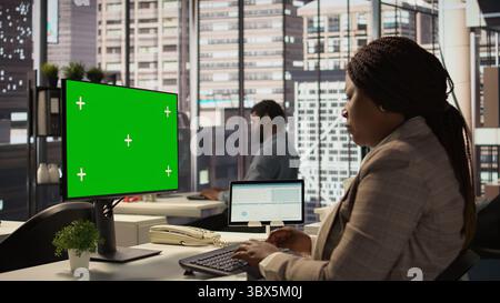 Female director seated at workstation with green screen on devices, working on solving administrative tasks for enterprise growth. Performance metrics to support project objectives. Camera B. Stock Photo