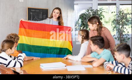 Schoolchildren sitting in classroom and listening to female teacher. Woman holding rainbow flag in hands and talking about minorities Stock Photo