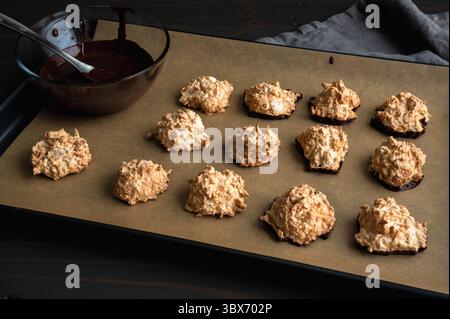 Macaroons Dipped in Dark Chocolate on a Sheet Pan: Coconut meringue drop cookies with a bowl of melted bittersweet chocolate on parchment paper Stock Photo