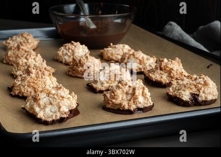 Macaroons Dipped in Dark Chocolate on a Sheet Pan: Coconut meringue drop cookies with a bowl of melted bittersweet chocolate on parchment paper Stock Photo