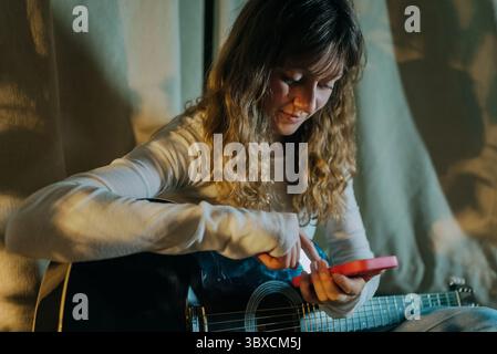 Young curly haired woman using smartphone while holding acoustic guitar, searching for chords or tabs online, enjoying music and technology at home Stock Photo