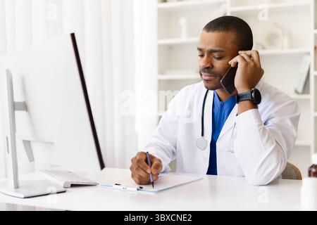 Focused African American doctor taking notes during phone conversation at work desk Stock Photo