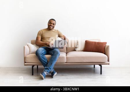 Smiling black man working remotely on laptop while sitting comfortably on sofa Stock Photo