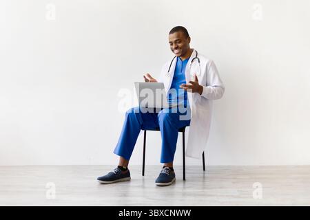 Cheerful black doctor using laptop during video call, gesturing with hands Stock Photo
