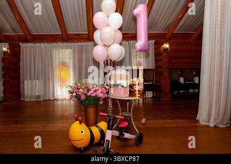 Festive indoor setup for a girl's first birthday featuring pink and white balloons, a number one foil balloon, flowers, gifts, a bee ride-on toy, a tr Stock Photo