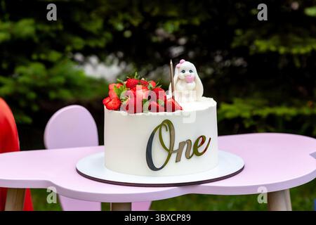 White frosted cake decorated with fresh strawberries and a cute bunny figurine, with the word One written on the front, placed on a pink high chair ou Stock Photo