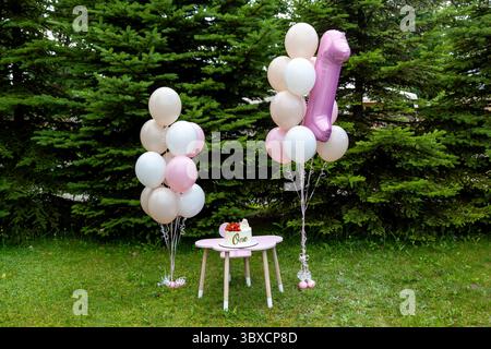 Adorable outdoor scene for a babys first birthday featuring a pink table with a One cake, surrounded by pastel balloons and a pink number one foil bal Stock Photo