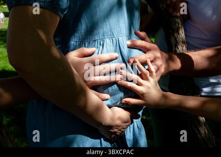 Close-up of multiple hands, including childrens, gently touching a pregnant womans belly outdoors, symbolizing love, unity, and anticipation of a new Stock Photo