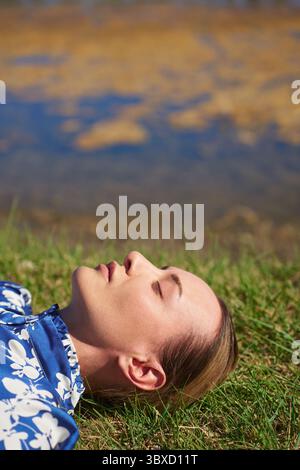 Serene woman relaxing and lying on a grass next to the pond Stock Photo