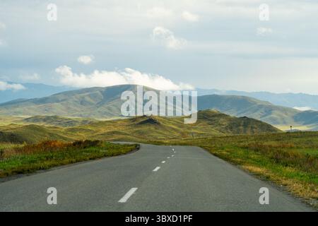 A long winding road stretches through a vast green mountainous Altai landscape under a partly cloudy sky. Stock Photo