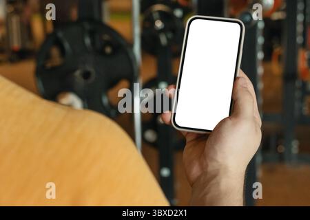 Athlete standing and displaying mobile phone with white blank screen in gym setting. Mock up. Space for design. Stock Photo