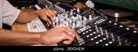 Young adult Black man adjusting audio mixing console in recording studio, hands operating sliders and knobs near computer keyboard and microphone, focused on sound engineering Stock Photo