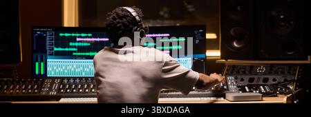 Black man young adult sitting at mixing console operating audio equipment in professional recording studio, wearing headphones and working on music production Stock Photo