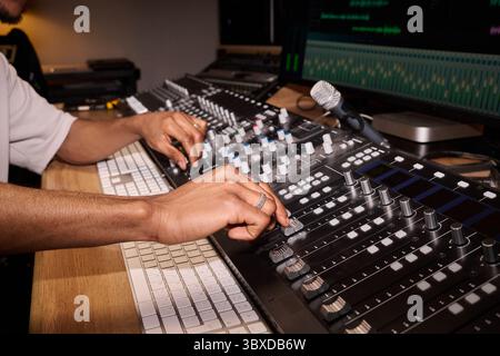 Young adult Black man adjusting audio mixing console in professional recording studio, hands operating multiple sliders and dials near computer keyboard and microphone Stock Photo