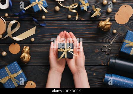 Hands hold out a blue gift box with gold ribbon surrounded by gift wrapping supplies on a blue wooden background Stock Photo