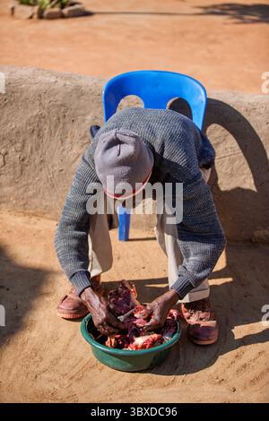 old african man sitting on the chair and cutting meat with a knife , village in Botswana in the yard doing daily chores Stock Photo